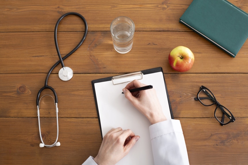 Recommendations and prescription from doctor. Woman in white coat writes on tablet at workplace, on wooden table with stethoscope, glasses, apple and glass of water, top view, copy space, cropped